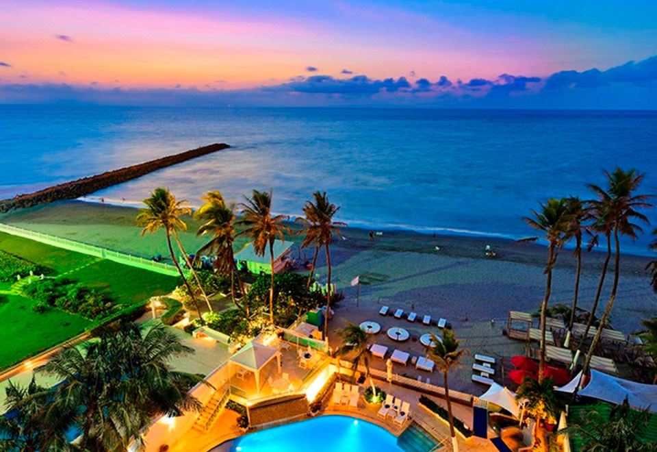 a large pool is surrounded by palm trees and a beachfront resort at dusk , with the ocean visible in the background at Hotel Dann Cartagena