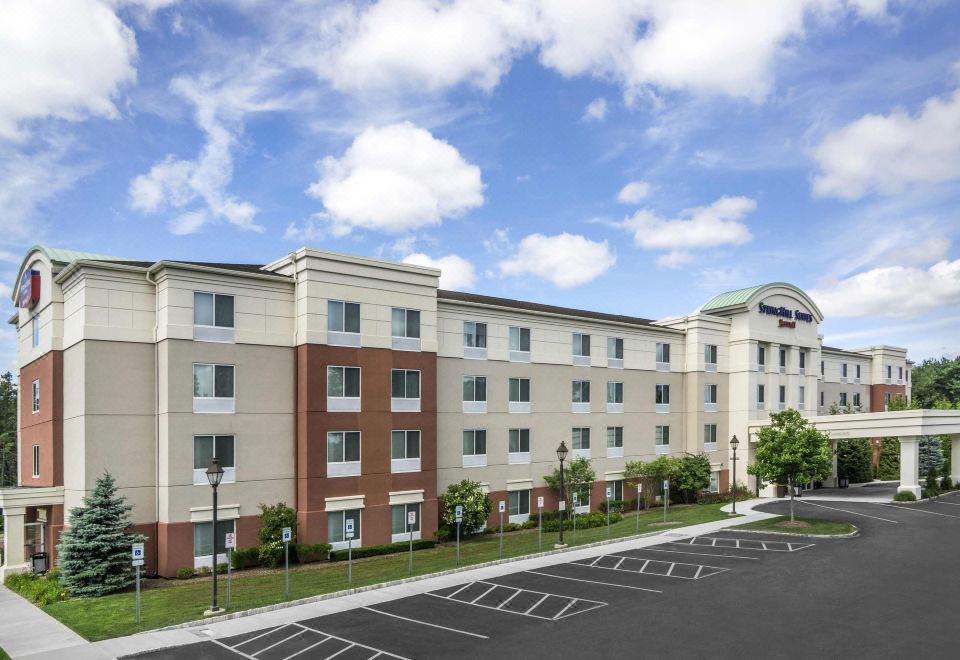 a large , multi - story hotel with a white and brown facade is surrounded by trees and has a parking lot in front at SpringHill Suites Long Island Brookhaven