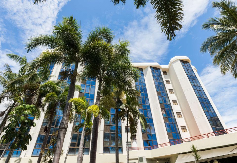 a tall building with blue windows and a red railing is surrounded by palm trees at Rydges Southbank Townsville, an EVT hotel