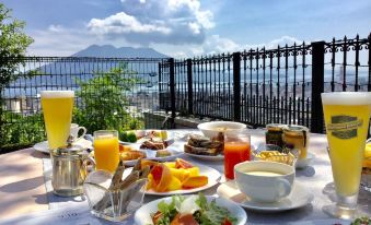 a table with a variety of breakfast items , including pastries , fruit , and beverages , is set up on a balcony overlooking the city at SHIROYAMA HOTEL kagoshima