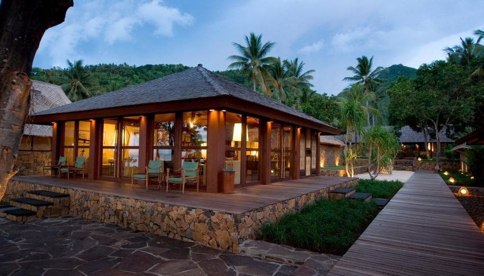 a modern wooden house with a stone terrace , surrounded by lush greenery and palm trees , at dusk at Jeeva Klui Resort