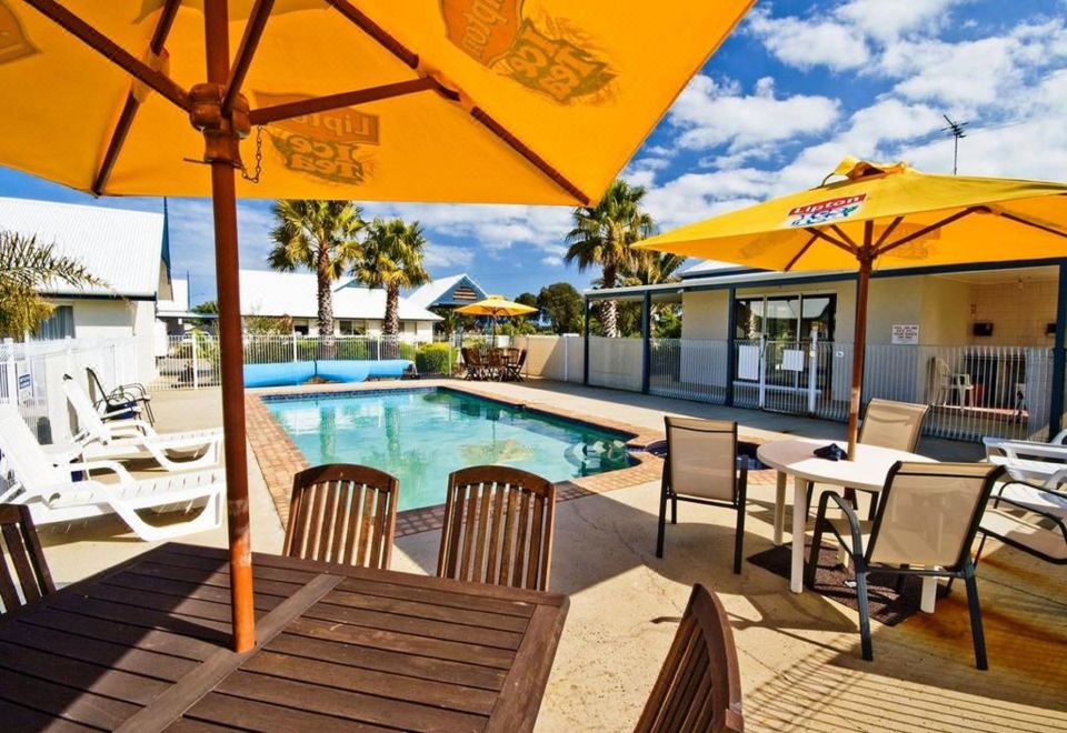 a sunny outdoor area with umbrellas , tables , and chairs near a swimming pool , surrounded by palm trees and buildings at Torquay Tropicana Motel