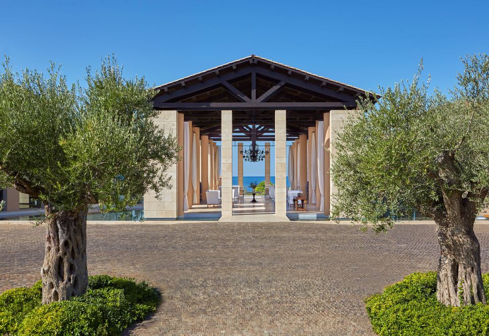 a large wooden building with columns and a terrace is surrounded by trees and grass at The Romanos, a Luxury Collection Resort, Costa Navarino