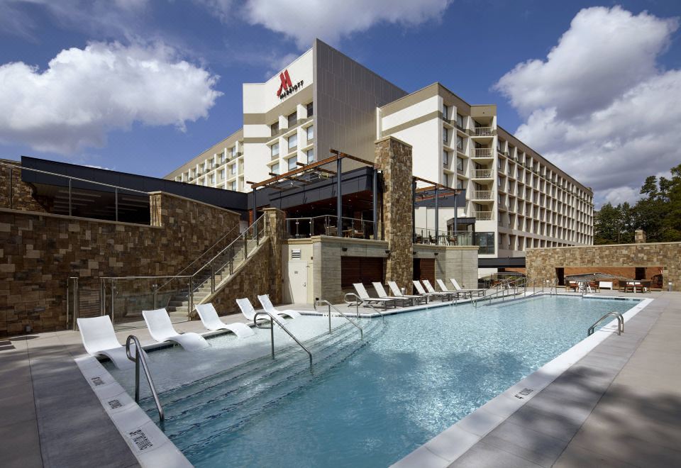 a large outdoor swimming pool surrounded by multiple buildings , with lounge chairs and umbrellas placed around the pool area at Raleigh Marriott Crabtree Valley