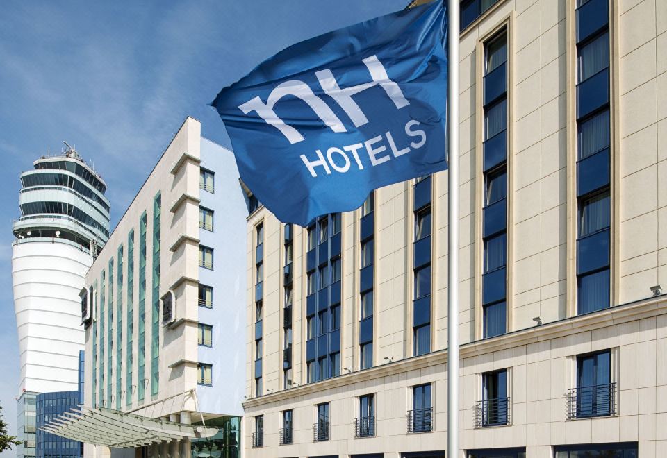 a large hotel with a blue and white flag flying in front of it , surrounded by cars parked outside at NH Vienna Airport Conference C