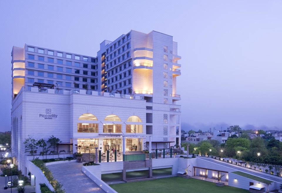 a modern hotel building surrounded by a well - maintained green lawn , with trees and a fountain in the background at Hyatt Centric Janakpuri New Delhi