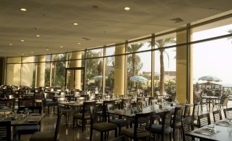 a large , empty restaurant with multiple dining tables and chairs , surrounded by windows that offer views of the outdoors at Dead Sea Spa Hotel