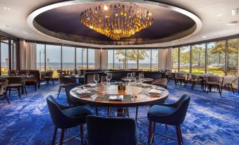 a modern dining room with a round table surrounded by chairs , a chandelier hanging from the ceiling , and large windows offering views of the ocean at The Grand Hotel Golf Resort & Spa, Autograph Collection