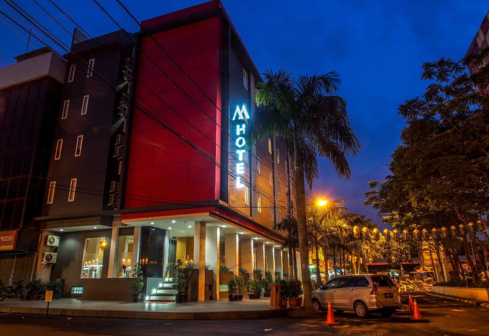 a modern hotel building with a red facade and a large sign on the front , surrounded by palm trees and street lights at night at M Hotel