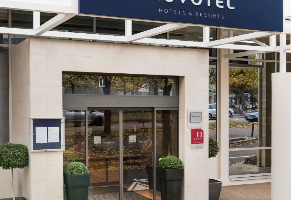 the entrance to the novotel hotel in europe , with the hotel 's name displayed above the door and two potted plants on either side of the at Novotel Chateau de Versailles