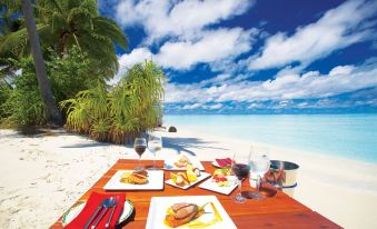 a table is set with plates of food and wine glasses on a beach , surrounded by palm trees and clouds at Filitheyo Island Resort