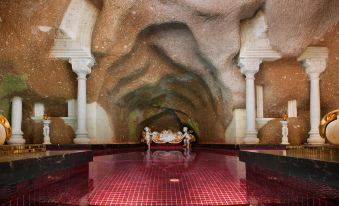 a large , empty pool with a cave - like design and two white columns is surrounded by red tiles at Sacred House