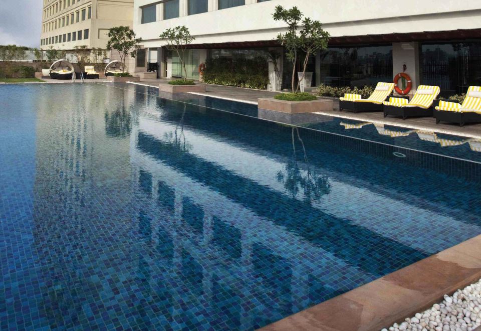 a large , blue tiled swimming pool surrounded by lounge chairs and trees , with a building in the background at Crowne Plaza New Delhi Mayur Vihar Noida