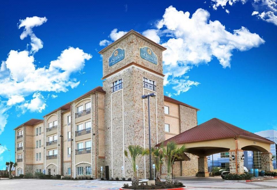 a large , modern hotel with a stone facade and a clock tower , surrounded by palm trees and cars in the parking lot at La Quinta Inn & Suites by Wyndham Dallas Grand Prairie South