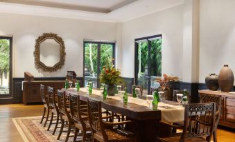 a long dining table set with green wine glasses and bottles , surrounded by chairs in a room with large windows at Plataran Heritage Borobudur Hotel
