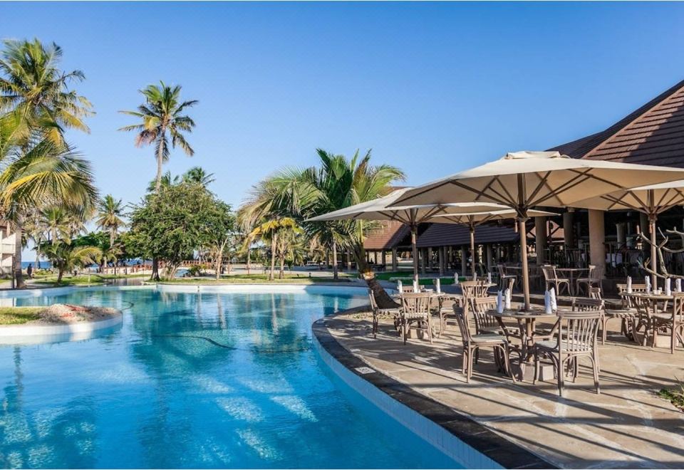 a resort with a large outdoor pool surrounded by palm trees , umbrellas , and tables and chairs at Amani Tiwi Beach Resort
