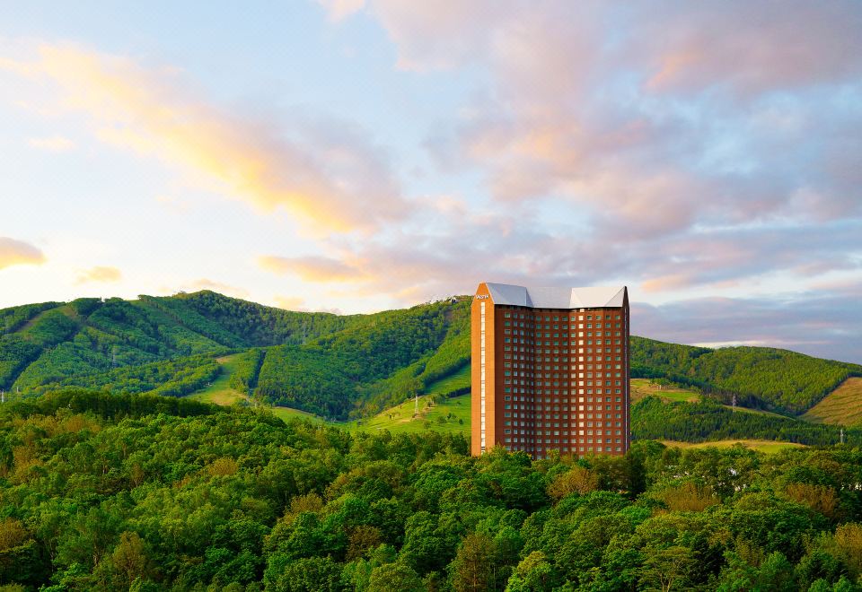 a tall building is nestled in a mountainous area with trees and hills in the background at The Westin Rusutsu Resort