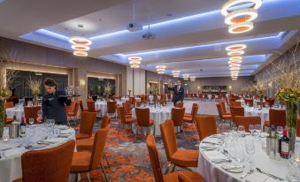 a large banquet hall with multiple round tables and chairs , orange chairs , and a man in the background at Clayton Hotel Chiswick