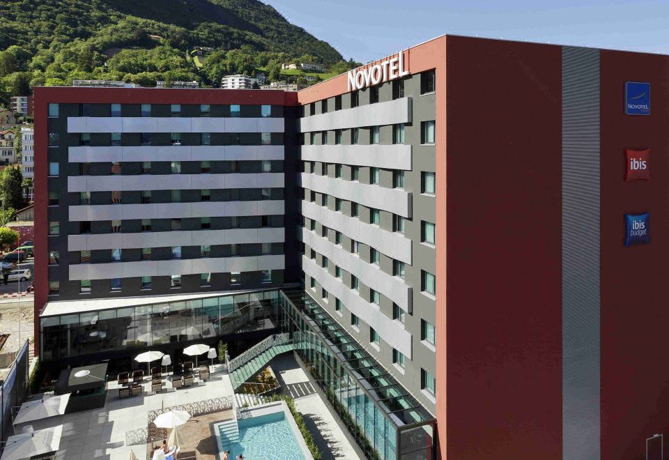 a modern hotel with a red and gray exterior , surrounded by trees and mountains , under a clear blue sky at Novotel Lugano Paradiso