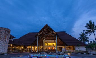 a large wooden building with a triangular roof is illuminated by blue lights , surrounded by water fountains and greenery at Amani Tiwi Beach Resort