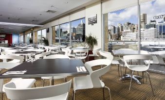 an empty restaurant with white tables and chairs , surrounded by large windows offering a view of the city at Ibis Sydney Darling Harbour