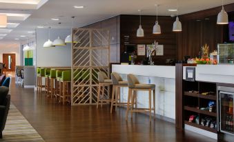 a modern bar area with wooden tables and chairs , a counter , and pendant lights hanging from the ceiling at Hampton by Hilton Exeter Airport