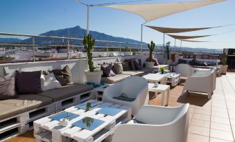 a rooftop patio with white furniture , including couches , chairs , and a dining table , surrounded by a beautiful view of the mountains at NH San Pedro