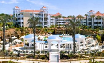 a large white building surrounded by palm trees and lush greenery , with a swimming pool in the background at Hilton Vilamoura As Cascatas Golf Resort & Spa