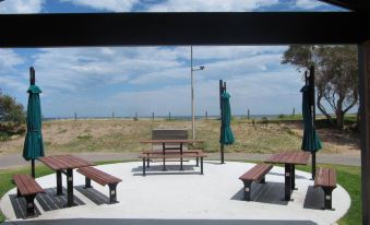 a picnic area with picnic tables , benches , and umbrellas under a shelter , overlooking a beach at Brighton Beachfront Holiday Park Adelaide