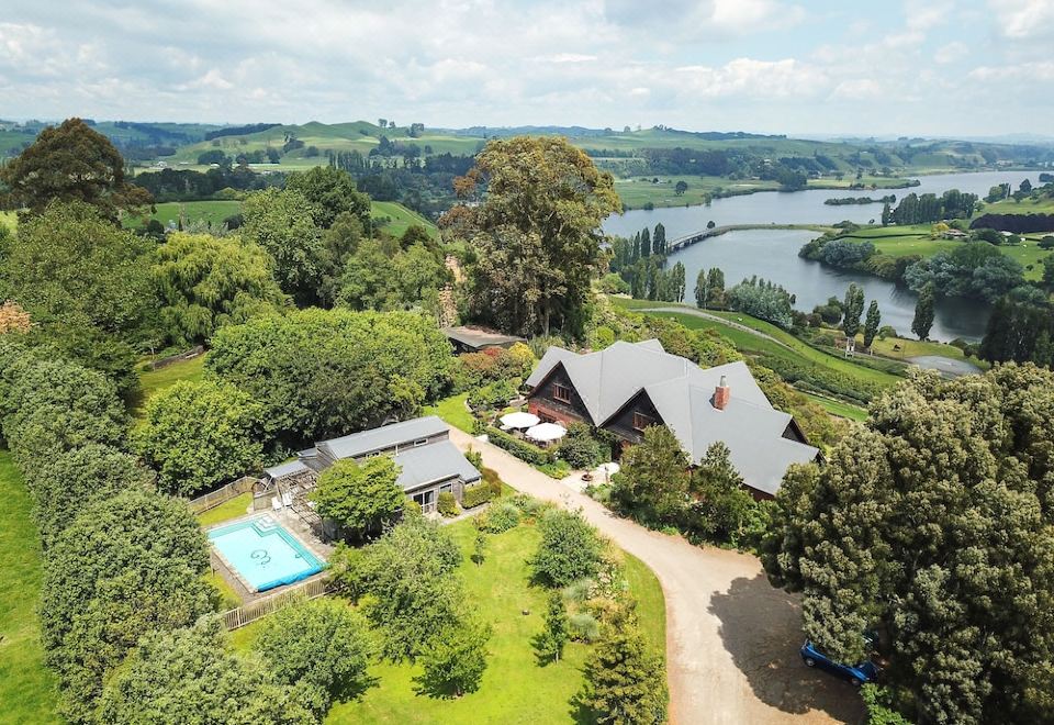 a large house surrounded by trees and a body of water , with a swimming pool in the foreground at Lake Karapiro Lodge