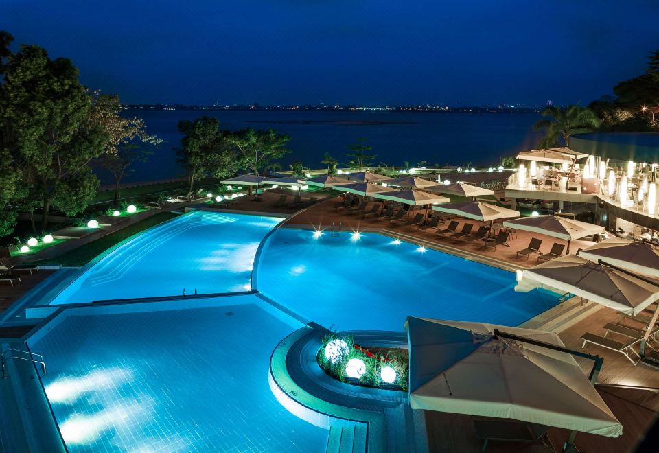 a large outdoor pool surrounded by lounge chairs and umbrellas , with a view of the ocean in the background at Radisson Blu M'Bamou Palace Hotel, Brazzaville