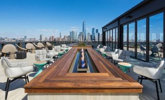 a rooftop terrace with wooden decking , white lounge chairs , and a central fire pit , overlooking a cityscape under a clear blue sky at Hyatt House Jersey City