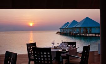 a wooden deck overlooking the ocean at sunset , with a dining table set up for a meal at Diamonds Athuruga Maldives Resort & SPA