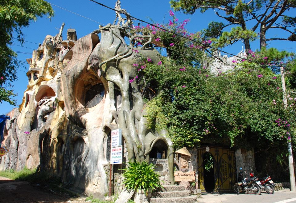 a building with a unique tree - like structure in the middle of the street , surrounded by cars and motorcycles at Crazy House