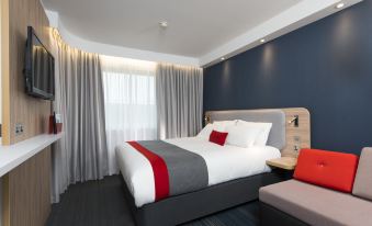 a modern hotel room with a large bed , gray curtains , and a red accent pillow at Holiday Inn Express Milton Keynes