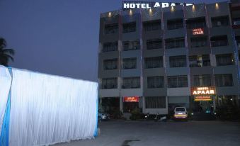 a tall hotel building is lit up at night , with a white building and car parked in front of it at Hotel Apaar