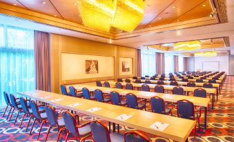 a large conference room with rows of tables and chairs , a chandelier hanging from the ceiling , and paintings on the walls at Leonardo Hotel Karlsruhe