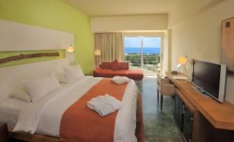 a modern hotel room with an orange bedspread , white pillows , and a large window offering a view of the ocean at E-Hotel Larnaca Resort & Spa