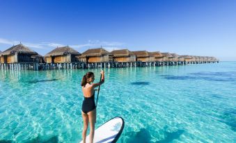 a woman standing on a surfboard in the middle of a body of water with houses in the background at Constance Moofushi