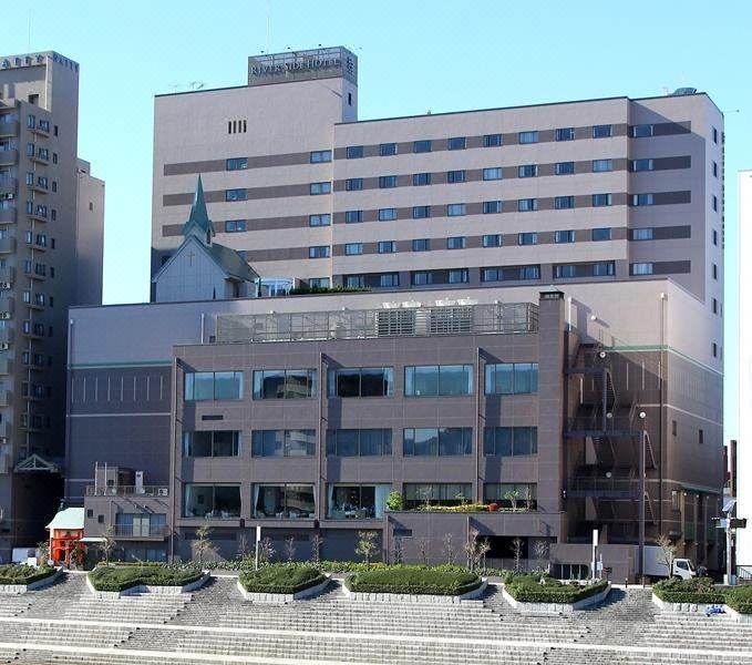 a modern building with multiple floors and large windows , surrounded by trees and other buildings at Numazu River Side Hotel