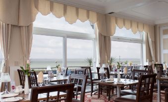a restaurant with wooden tables and chairs , white curtains , and a view of the ocean at Best Western New Holmwood Hotel