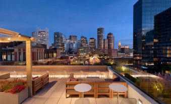 a rooftop patio with wooden furniture and a city skyline in the background , lit up at night at Yotel Boston