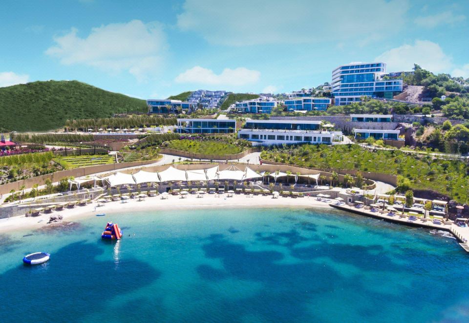 a beach resort with a white sand and blue water , surrounded by buildings and palm trees at Le Méridien Bodrum Beach Resort