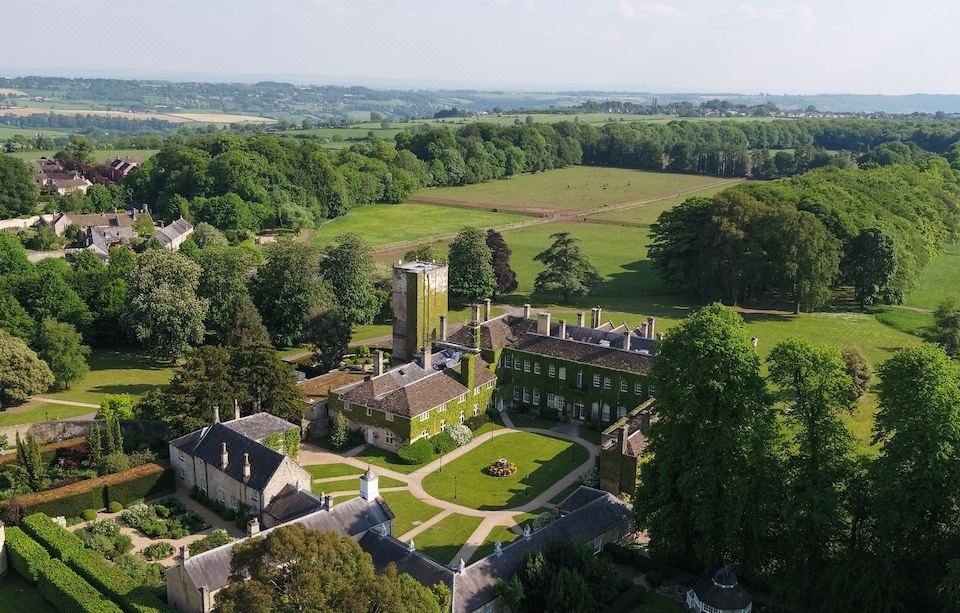 aerial view of a large green and white building surrounded by grass , trees , and grass - covered fields at Lucknam Park Hotel