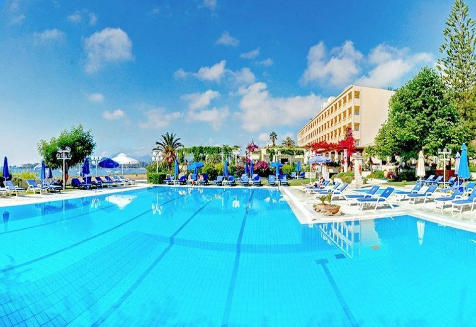 a large outdoor swimming pool surrounded by lounge chairs and umbrellas , with a hotel in the background at Corfu Palace Hotel
