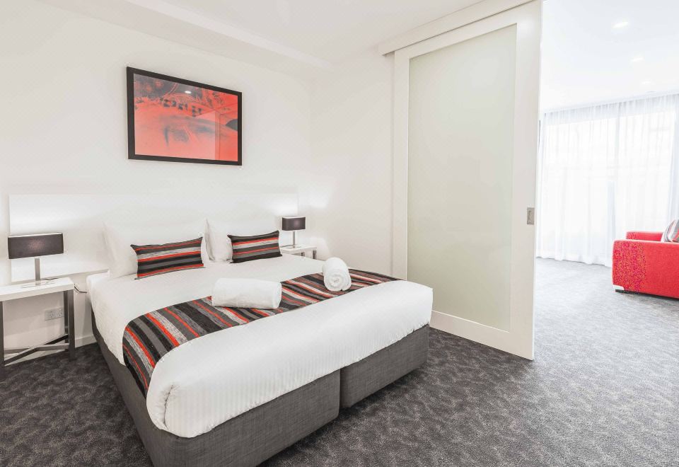 a large bed with white linens and a red and black striped blanket is in a hotel room at City Edge Box Hill Apartment Hotel
