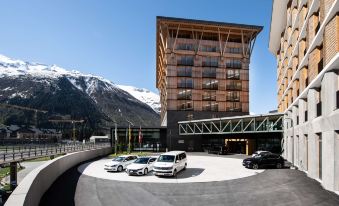 a parking lot with several cars parked in front of a tall building with mountains in the background at Radisson Blu Hotel Reussen Andermatt