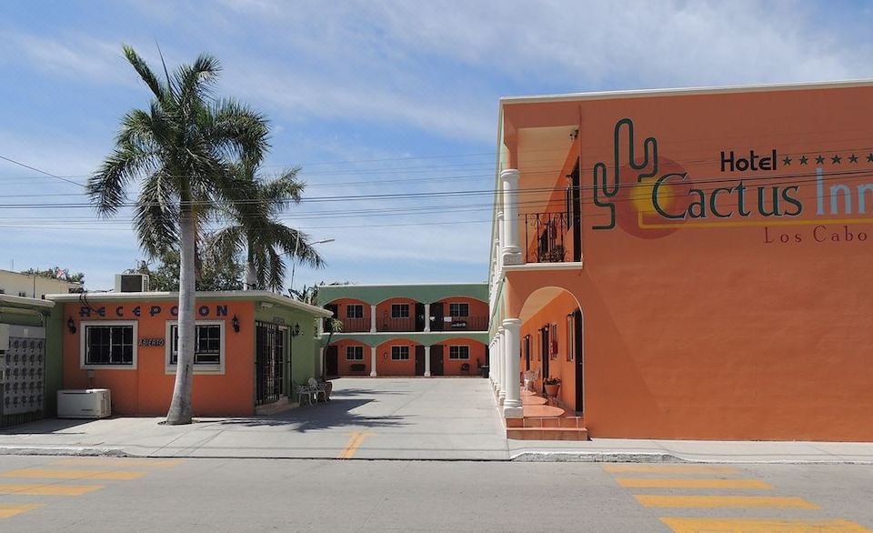 a red and orange building with a cactus on the side is surrounded by palm trees at Cactus Inn Los Cabos