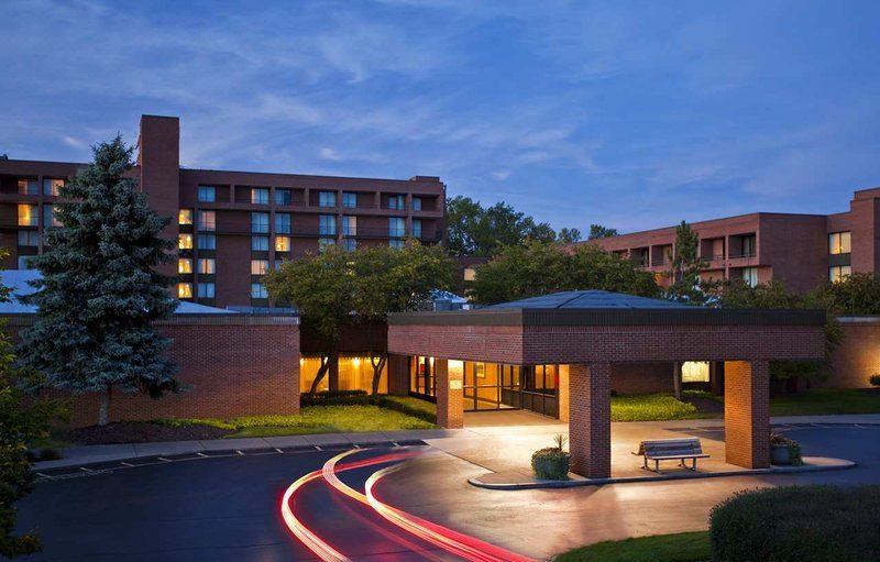 a modern hotel with a lit - up entrance and parking lot , set against a backdrop of trees at dusk at DoubleTree by Hilton Hotel Syracuse