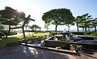 a wooden deck with a water fountain , surrounded by trees and grass , and the sun shining brightly in the background at Paradise Hotel Busan